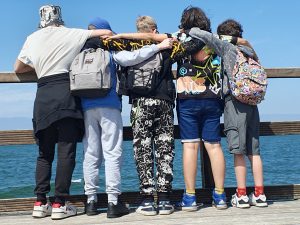 Pięcioro uczniów stoi na drewnianym pomoście i patrzy się w stronę morza znajdującego się przed nimi. Chłopcy ustawieni są tyłem do obiektywu i obejmują się ramionami.