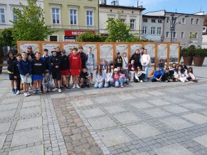 Uczniowie biorący udział w wycieczce pozują do wspólnego zdjęcia na tle ustawionych obok siebie dziesięciu tablic zawierających informacje historyczne. Część dzieci stoi, inne siedzą na chodniku przed tablicami.