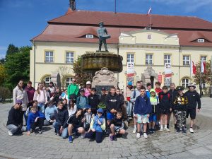 Uczniowie biorący udział w wycieczce pozują do grupowego zdjęcia na tle fontanny ustawionej na rynku w Darłowie. Fontanna na cokole ma ustawiony posąg żeglarza trzymającego w dłoniach łódź. W tle widać okazały budynek przed którym umieszczono polskie flagi.