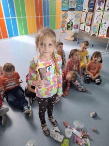 Uczennica oddziału przedszkolnego z blond warkoczem, ubrana w kolorową bluzkę w kształty przypominające chmurki i ciemne legginsy z truskawkami, stoi na tle kolorowych szafek. Trzyma w ręku zużyte opakowanie po batoniku. Za nią siedzi grupa dzieci na podłodze, a obok leżą różne odpady, m.in. aluminiowa folia, plastikowe opakowania, puste kubeczki i tubki. W tle widać plansze z dziecięcymi rysunkami przedstawiającymi globusy.