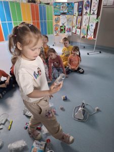 Uczennica oddziału przedszkolnego w jasnym ubraniu trzyma w ręku przezroczystą plastikową butelkę, stojąc przed grupą dzieci siedzących na podłodze na szkolnym korytarzu. Na podłodze rozsypane są różne odpady, a w tle widać kolorowe szafki i plansze z dziecięcymi rysunkami globusów.