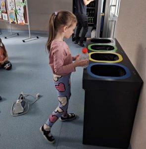 Uczennica oddziału przedszkolnego w różowej bluzce i legginsach z kolorowymi sercami stoi przy zestawie pojemników do segregacji odpadów, trzymając plastikową butelkę z czerwoną nakrętką. W tle widoczna inna dziewczynka siedząca na podłodze oraz plansze z rysunkami dzieci.