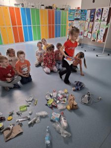 Uczennica oddziału przedszkolnego ubrana w czerwoną koszulkę podnosi książkę z podłogi, na której leżą różnorodne odpady. Za nią siedzi grupa dzieci, a w tle widać kolorowe szafki oraz plansze z dziecięcymi pracami plastycznymi, przedstawiającymi globusy.