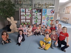 Uczniowie oddziału przedszkolnego pozują do zdjęcia na szkolnym korytarzu na tle plansz z pracami plastycznymi i informacjami o ochronie środowiska z okazji Dnia Ziemi. Obok planszy stoi dekoracyjny drewniany królik, a za dziećmi widać gablotę z pucharami.