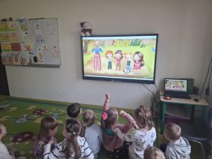 Grupa dzieci z oddziału przedszkolnego siedzi na kolorowym dywanie w swojej sali, zwrócona w stronę dużego monitora zawieszonego na ścianie. Na ekranie wyświetlony jest film animowany przedstawiający nauczycielkę i czworo dzieci stojących w parku. Obraz ten jest również widoczny na mniejszym ekranie laptopa stojącego na stoliku po prawej stronie. Obok monitora znajduje się biała tablica z przyczepionymi kartkami edukacyjnymi i rysunkami. Nad monitorem leży pluszowy miś. Dzieci patrzą uważnie w stronę ekranu, jedno z nich unosi rękę.