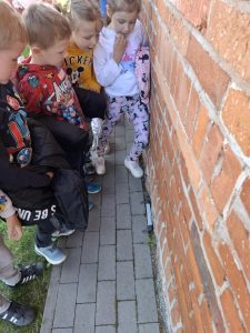 Grupa pięciorga dzieci z oddziału przedszkolnego stoi na zewnątrz przy czerwonej ceglanej ścianie budynku, patrząc z zainteresowaniem na termometr przymocowany nisko do ściany. Dzieci stoją na wąskiej ścieżce z szarej kostki brukowej, a za nimi widać kawałek trawnika. Wszystkie dzieci są ubrane w kolorowe bluzy z motywami bajkowymi – w tym Avengers, Mickey Mouse i Minnie Mouse. Jedno z dzieci trzyma kurtkę, inne ma rękę przy ustach w zamyśleniu. Panuje atmosfera ciekawości i zaangażowania.