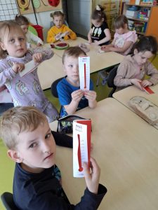 Grupa uczniów z oddziału przedszkolnego siedzi przy stolikach w swojej sali i bierze udział w zajęciach edukacyjnych. Dwoje dzieci na pierwszym planie pokazuje własnoręcznie wykonane termometry z papieru z napisem 'Mały Odkrywca'. Termometry mają narysowaną skalę od 0 do 20 i czerwoną wskazówkę imitującą poziom temperatury. Pozostałe dzieci w tle również pracują nad swoimi projektami, składając i ozdabiając papierowe termometry. Na stole leżą różne materiały plastyczne, a w tle widać kolorowe półki i plansze edukacyjne.