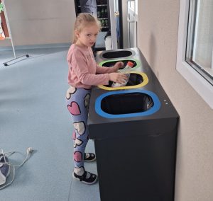 Uczennica oddziału przedszkolnego w różowej bluzce i legginsach w kolorowe serca wrzuca plastikową butelkę do jednego z kolorowych pojemników do segregacji odpadów w szkole. Stoi obok pojemników oznaczonych kolorami: niebieskim, żółtym i zielonym