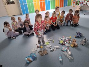 Uczennica oddziału przedszkolnego podnosi odpadek z podłogi na szkolnym korytarzu, gdzie przed nią leży rozrzucone wiele różnych śmieci. W tle siedzi grupa dzieci na tle kolorowych szafek i prac plastycznych zawieszonych na tablicy.