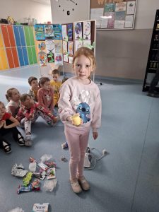 Na zdjęciu widać grupę dzieci oddziału przedszkolnego. Na pierwszym planie stoi dziewczynka z blond włosami zaplecionymi w warkocz, ubrana w kolorową bluzkę w serduszka i ciemne legginsy z nadrukiem. Trzyma w ręce pustą tubkę po produkcie spożywczym, przygotowaną do sortowania odpadów. Za nią siedzi grupa dzieci na podłodze, a przed nimi leży rozrzucone różnorodne opakowania – plastikowe, aluminiowe i papierowe. W tle widać kolorowe szafki (niebieskie, zielone, pomarańczowe i żółte) oraz tablice z pracami plastycznymi dzieci, przedstawiającymi rysunki globusów.