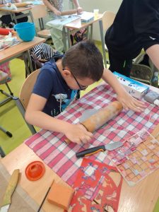 Uczeń klasy drugiej siedzi przy szkolnej ławce zakrytej ścierką w czerwoną kratę. Na stole leżą szablony i przyrządy do wykonania pracy plastycznej. Chłopiec energicznie przyciska obiema dłońmi wałek do szarej masy plastycznej.