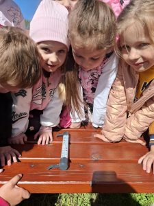 Grupa dzieci z oddziału przedszkolnego jest pochylona nad drewnianą ławką na świeżym powietrzu, na której leży analogowy termometr rurkowy w plastikowej obudowie. Dzieci z zaciekawieniem obserwują temperaturę, niektóre z nich dotykają ławki rękami. Dziecko po lewej stronie ma jasnoróżową czapkę i kurtkę z napisem 'Athletics', obok stoi dziewczynka w bluzie z nadrukiem Minnie Mouse. Po prawej stronie widoczna jest dziewczynka w jasnobeżowej pikowanej kurtce oraz inne dzieci w tle. Promienie słońca padają na ich twarze i na drewnianą powierzchnię ławki, a w dolnej części zdjęcia widoczny jest cień fotografa.