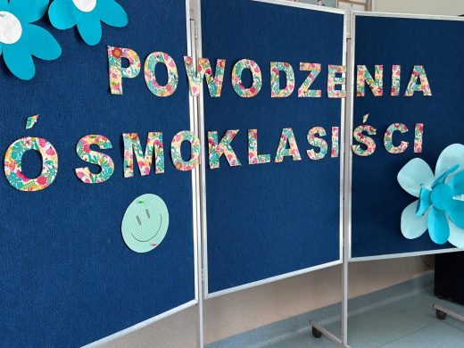 Kolorowa dekoracja szkolna z napisem 'Powodzenia ósmoklasiści' ułożonym z liter pokrytych wzorem kwiatowym na granatowym tle parawanu. Obok znajdują się duże papierowe kwiaty i uśmiechnięta buźka z papieru.