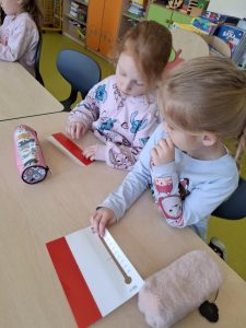Dwie dziewczynki z oddziału przedszkolnego siedzą przy stoliku w swojej sali i pracują nad papierowymi termometrami edukacyjnymi. Obie ubrane są w bluzy z dziecięcymi motywami — jedna z postacią Stitcha, druga z bohaterkami bajki. Dziewczynki skupione są na zadaniu – jedna z nich palcem przesuwa czerwoną wskazówkę na papierowym termometrze z podziałką od 0 do 20 stopni. Na stole leżą ich piórniki, w tym jeden pluszowy. W tle widoczne są kolorowe szafki, półki z książkami i pomoce edukacyjne