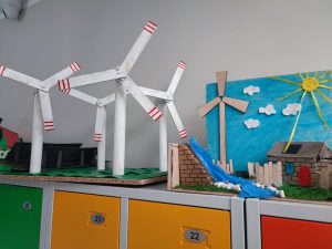 Zbliżenie na kilka makiet ustawionych na kolorowych szafkach. Makiety przedstawiają m.in. wiatraki, domki z panelami słonecznymi, elektrownię wodną i słoneczny krajobraz z niebem, chmurkami i promieniującym słońcem wykonanym z żółtej wstążki.
