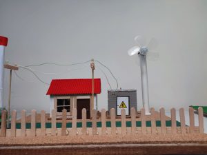 Zdjęcie miniaturowej makiety domu z czerwonym dachem, ogrodzeniem z patyczków i turbiną wiatrową z przezroczystymi łopatami. W tle linie energetyczne i skrzynka elektryczna.