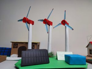 Zdjęcie makiety przedstawiającej trzy duże wiatraki z czerwonymi gondolami i niebieskimi łopatami. Przed nimi czarny panel słoneczny i niebieskie pudełko. Z boku znajduje się kartka z napisem „Odnawialne źródła energii”.