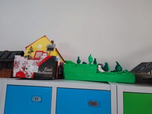 Zdjęcie modelu przedstawiającego dom z żółtą fasadą i czerwonym dachem, stojącego na kartonowym pudełku. Obok znajduje się zielony model ogrodu lub pola z roślinami wykonanymi z zielonych materiałów.