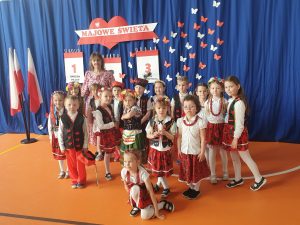 Uczniowie oddziału przedszkolnego pozują do wspólnego zdjęcia wraz ze swoją wychowawczynią. Dzieci ubrane są w stroje krakowiaków. W tle na niebieskim materiale zawieszona jest dekoracja w białym i czerwonym kolorze - napis "MAJOWE ŚWIETA" oraz trzy dekoracje przedstawiające kartki z kalendarza ponumerowane od 1 do 3 z wypisanymi pod nimi nazwami świąt. całość uzupełniają białe i czerwone motyle wycięte z papieru.
