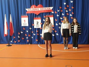 W centralnej części zdjęcia widoczna jest uczennica klasy piątej trzymająca przed sobą mikrofon. za nią stoją jej dwie koleżanki. W tle na niebieskim tle zawieszona jest dekoracja utrzymana w białym i czerwonym kolorze - - napis "MAJOWE ŚWIETA" oraz trzy dekoracje przedstawiające kartki z kalendarza ponumerowane od 1 do 3 z wypisanymi pod nimi nazwami świąt. całość uzupełniają białe i czerwone motyle wycięte z papieru.