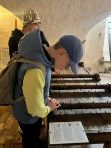 Uczeń pochyla się nad urządzeniem do tortur znajdującym się w sali muzealnej. Chłopiec przygląda się opisowi umieszczonemu obok maszyny.