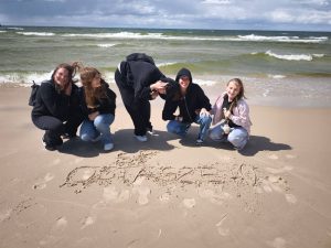 Pięć uczennic pozuje do wspólnego zdjęcia na plaży. Cztery z nich kucają na piasku, a dziewczyna stojąca po środku pochyla się bokiem zasłaniając częściowo swoją twarz. Na piasku przed uczennicami widoczny jest napis "SO OSTASZEWO".