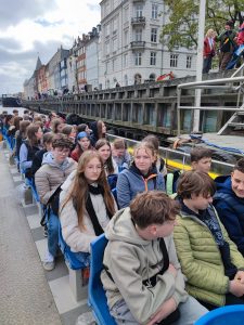 Uczniowie bioracy udział w wycieczce siedzą w trzy osobowych rzędach utworzonych z niebieskich, plastikowych foteli na pokładzie łodzi. W tle widoczne są zabudowania portowego miasta obok, którego stacjonuje łódź.
