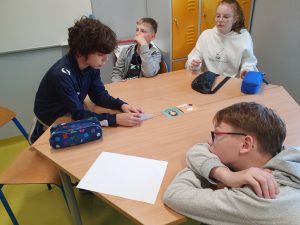 Czworo dzieci siedzi przy szkolnym, sześciokątnym stole w klasie. Dwóch chłopców po lewej stronie patrzy na kartkę trzymaną przez jednego z nich. Dziewczynka w białej bluzie z nadrukiem królika siedzi po prawej stronie i trzyma piórnik. Na stole leży kilka przedmiotów: piórniki, kartka papieru i kolorowa karta z rysunkiem postaci. Dziecko na pierwszym planie, z okularami i jasną bluzą, opiera się głową o rękę, spoglądając w stronę kartki. W tle widać żółte szafki szkolne i tablicę.