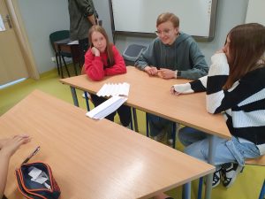 Trzy uczennice klasy szóstej siedzą przy wspólnym stole. W lekkim oddaleniu od nich ustawiona jest druga ławka. Pomiędzy ławkami leży most wykonany ze zgiętej w harmonijkę kartki papieru. Dziewczynki patrzą w jego stronę.