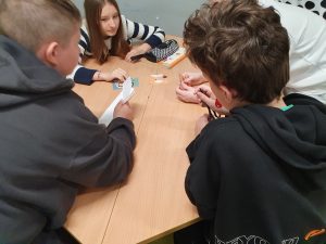 Grupa pięciorga dzieci siedzi przy sześciokątnym stole w klasie. Dwoje chłopców w ciemnych bluzach siedzi tyłem do aparatu – jeden z nich trzyma kartkę papieru. Naprzeciwko nich siedzi dziewczynka w pasiastym swetrze oraz dwóch uczniów, częściowo przysłoniętych. Na stole leżą kolorowe karty z ilustracjami postaci. W tle widać fragment ściany z czarno-białą grafiką i książkę leżącą na stole.