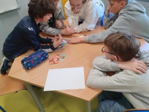 Pięcioro dzieci siedzi przy sześciokątnym stole w sali lekcyjnej. Są skupione na wspólnej aktywności — oglądają i omawiają kolorowe ilustracje z postaciami, które leżą na stole. Dzieci mają na sobie bluzy i dresy, a na stole leży również piórnik i kartka papieru. W tle widać fragment białej tablicy i kolorowe szafki. Scena przedstawia zaangażowaną pracę grupową.