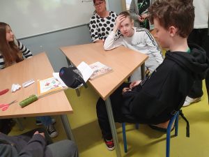 Grupa uczniów siedzi w klasie przy połączonych ławkach. Na stole leżą podręczniki, piórnik, kartki papieru, nożyczki, sznurek i papierowe ozdoby. Jeden chłopiec opiera głowę na ręce, patrząc w stronę aparatu, inny siedzi tyłem. Po lewej stronie widać dziewczynę w pasiastym swetrze. W tle stoi nauczycielka w okularach i białej bluzce w czarne grochy, patrzy na uczniów. Tło stanowi biała tablica i jasnozielona podłoga. Scena ukazuje zajęcia w klasie z budowania relacji w grupie.