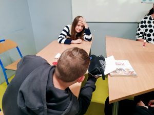 "Troje uczniów siedzi przy dwóch połączonych ławkach w klasie. Dziewczyna w pasiastym swetrze opiera głowę na ręce i patrzy w kierunku chłopca, który pochyla się nad piórnikiem i zeszytem. Na stole leży podręcznik do języka polskiego 'Czas na start'. W tle biała tablica i niebieska ściana."