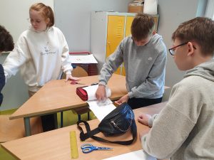 Czworo uczniów pracuje wspólnie przy ustawionych w literę L ławkach w klasie. Dziewczyna w białej bluzie z napisem ‘Enjoy every moment’ i rudych włosach związanych w kucyk spogląda w lewo, opierając rękę na ławce. Dwóch chłopców w szarych bluzach pochylonych nad otwartym zeszytem i przyborami – nożyczkami, linijką, piórnikiem i czarną nerką – pracuje nad zadaniem. W tle żółta szafka i rysunki na kartkach papieru.