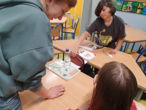 Troje uczniów tworzy z kartki papieru most pomiędzy dwoma oddalonymi od siebie ławkami. Most przytrzymywany jest na ławkach za pomocą szkolnego podręcznika i zeszytu. Chłopiec widoczny na zdjęciu ustawia na moście piórnik.