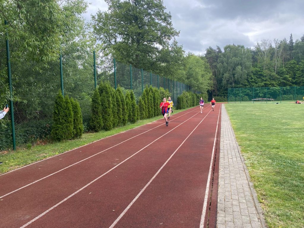 Na bieżni z czteroma torami biegną cztery zawodniczki. Na przodzie biegnie uczennica naszej szkoły. W tle widać zieloną murawę i drzewa okalające boisko sportowe.