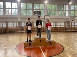 Na sali gimnastycznej ustawione jest podium z miejscami ponumerowanymi od 1 do 3. Na każdym miejscu podium stoi chłopak z zawieszonym na szyi medalem. Chłopcy trzymają w dłoniach przed sobą dyplomy. Na miejscu podpisanym numerem 3 stoi uczeń naszej szkoły.