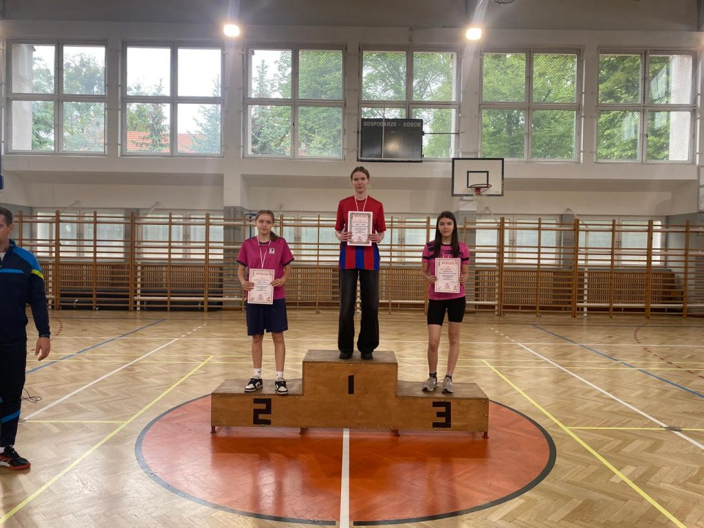 Na sali gimnastycznej ustawione jest podium z miejscami ponumerowanymi od 1 do 3. Na każdym miejscu podium stoi dziewczyna z zawieszonym na szyi medalem. Dziewczyny trzymają w dłoniach przed sobą dyplomy. Na miejscu podpisanym numerem 1 stoi uczennica naszej szkoły.