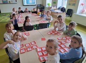 Uczniowie oddziału przedszkolnego siedzą przy dwóch dużych stołach. Dzieci patrzą w stronę obiektywu. Przed nimi leżą odręcznie wykonane znaki zakazu palenia przyklejone na czerwonych kwadratowych kartkach papieru.