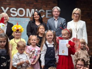 Część uczestników konkursu wraz ze swoimi opiekunami i przedstawicielami z gminy pozują do wspólnego zdjęcia stojąc na ścianie wykonanej z cegieł z napisem "Gmina Łysomice". Jedna z dziewczynek ubrana na czerwono trzyma przed sobą otrzymany dyplom. Zdjęcie wykonane w zbliżeniu.