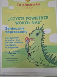 Plakat informacyjny o programie edukacyjnym dla przedszkoli. Na jasnozielonym tle znajduje się kolorowa ilustracja uśmiechniętego zielonego dinozaura trzymającego bukiet kwiatów. W tle widać białe chmurki i pagórkowaty krajobraz. Treść plakatu: Ta placówka realizuje Program Przedszkolnej Edukacji Antytytoniowej pt. "Czyste powietrze wokół nas" Serdecznie zapraszamy przedszkolaki w wieku 6 lat oraz ich rodziców do udziału w programie! Dyrekcja i pracownicy przedszkola oraz Państwowy Powiatowy Inspektor Sanitarny w Toruniu Na dole znajduje się adres strony internetowej: www.torun.psse.gov.pl Plakat ma charakter edukacyjno-promocyjny i jest skierowany do dzieci w wieku przedszkolnym oraz ich rodziców, zachęcając do udziału w programie antynikotynowym.