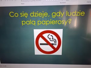 Zdjęcie przedstawia slajd z prezentacji multimedialnej wyświetlonej na monitorze komputera. Tytuł slajdu, zapisany dużymi żółtymi literami na ciemnym tle, brzmi: „Co się dzieje, gdy ludzie palą papierosy?”. Pod tytułem zamieszczony jest znak zakazu palenia.