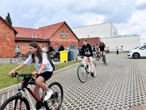 Dziewczęta na rowerach wyjeżdżają ze szkolnego parkingu wyłożonego kostką brukową. W tle czerwone budynki szkoły z cegły oraz nowoczesny biały budynek. Pogoda pochmurna, kilka osób stoi w tle przy stojakach na rowery.