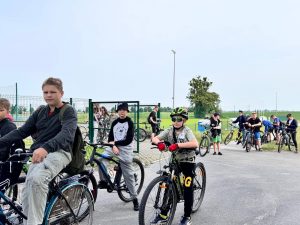 Uczniowie na rowerach wyjeżdżają poza bramę szkolnego parkingu przygotowując się do rowerowego rajdu. W tle widać boisku i zieloną murawę. Niebo jest zamglone.