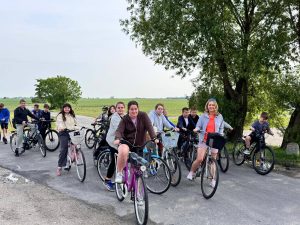 Grupa uczniów i nauczycielka na rowerach na wiejskiej drodze, otoczonej polami i drzewami. Wszyscy uśmiechnięci, gotowi do dalszej jazdy. Drzewo po prawej daje cień, a w tle rozciągają się zielone pola pod zachmurzonym niebem.