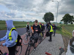 Uczniowie biorący udział w rajdzie zatrzymują się na postoju przy asfaltowej drodze. W oddali widoczne są pojedyncze drzewo. Niebo jest zachmurzone.