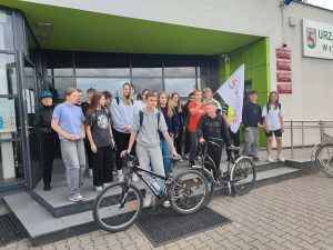 Uczniowie pozują do wspólnego zdjęcia przed budynkiem Urzędu Gminy Łysomice. Dwoje uczniów stojących z przodu opiera się o rowery. Chłopiec za nimi trzyma w dłoniach flagę gminy Łysomice.