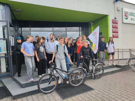 Uczniowie pozują do wspólnego zdjęcia przed budynkiem Urzędu Gminy Łysomice. Dwoje uczniów stojących z przodu opiera się o rowery. Chłopiec za nimi trzyma w dłoniach flagę gminy Łysomice.