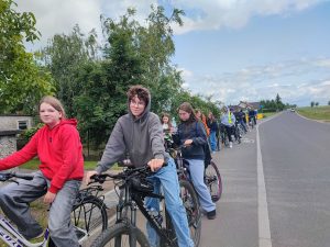 Uczniowie biorący udział w rajdzie rowerowym stoją na ścieżce oznaczonej jako droga dla rowerów przy asfaltowej drodze. po prawej stronie ścieżki widoczne są drzewa, a na lewo pola.