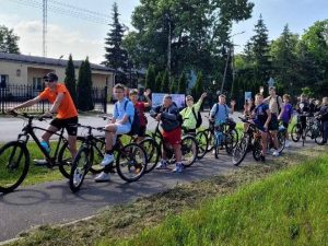 Grupa uczniów na rowerach ustawiona w rzędzie na ścieżce rowerowej przy drodze. Dzieci i młodzież w różnym wieku uśmiechają się i machają do obiektywu. W tle budynek z flagą i ogrodzeniem, drzewa iglaste i liściaste. Pogoda słoneczna.